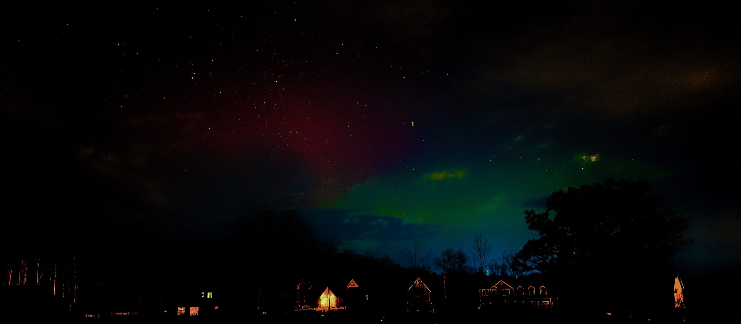 Wide panorama of green and red aurora over a row of houses with lit windows.